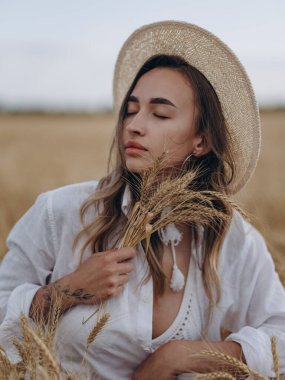 Hasır şapkalı, beyaz gömlekli, dinlenen ve gevşeyen genç bir kadın. Elinde olgun buğday kulaklarıyla tarlada dinleniyor. Huzurlu kırsal yaz manzarası, doğal güzellik. Yakın plan. Portre.