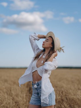 Hasır şapkalı, beyaz gömlekli, sütyenli, dinlenen, gözleri kapalı, elinde olgun buğday kulakları olan genç bir kadın. Huzurlu kırsal yaz manzarası, doğal güzellik.