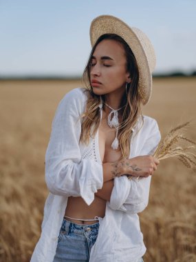 Hasır şapkalı, beyaz gömlekli, sütyenli, dinlenen, gözleri kapalı, elinde olgun buğday kulakları olan genç bir kadın. Huzurlu kırsal yaz manzarası, doğal güzellik. Yakın plan..