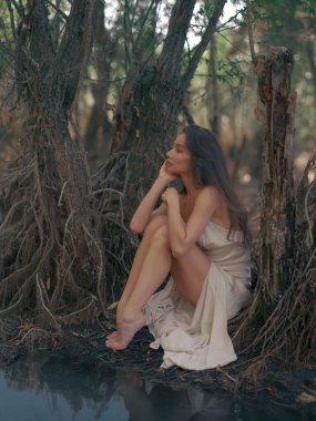 Beyaz elbiseli genç kadın, ağaç kökleri ile çevrili ve suya yansıyan orman gölünün kenarında yalınayak oturuyor. Doğadaki sakin atmosfer. Peri masalı, büyülü, gizemli ve mistik orman. Yakın plan..