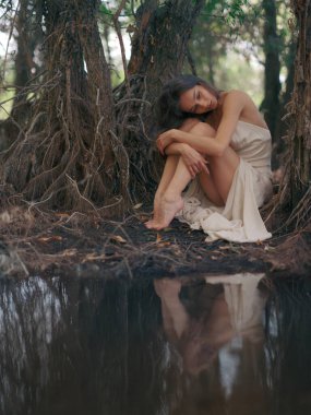 Beyaz elbiseli genç kadın, ağaç kökleri ile çevrili ve suya yansıyan orman gölünün kenarında yalınayak oturuyor. Doğadaki sakin atmosfer. Peri masalı, büyülü, gizemli ve mistik orman. Yakın plan..