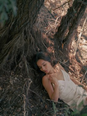 Beyaz elbiseli genç esmer kadın yere uzanıyor ve yaşlı ağaç kökleriyle çevrili ormanda gözleri kapalı dinleniyor. Sakin atmosfer, dinlenme, dinlenme ve meditasyon. Üst görünüm.