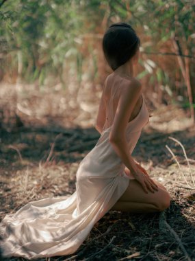 Beyaz elbiseli genç esmer kadın ormanda yerde oturuyor ve rahatlıyor. Sakin atmosfer, dinlenme, rahatlama ve doğada meditasyon. Yan görünüm.