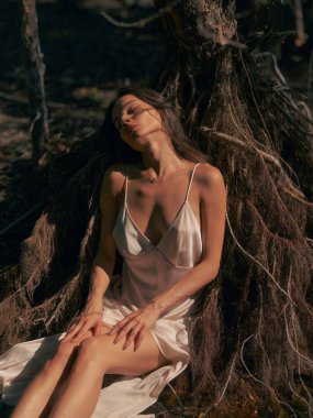 Genç esmer, beyaz elbiseli bir kadın yerde oturuyor ve yaşlı ağaç kökleriyle çevrili ormanda gözleri kapalı dinleniyor. Sakin bir atmosfer, dinlenme ve doğada meditasyon..
