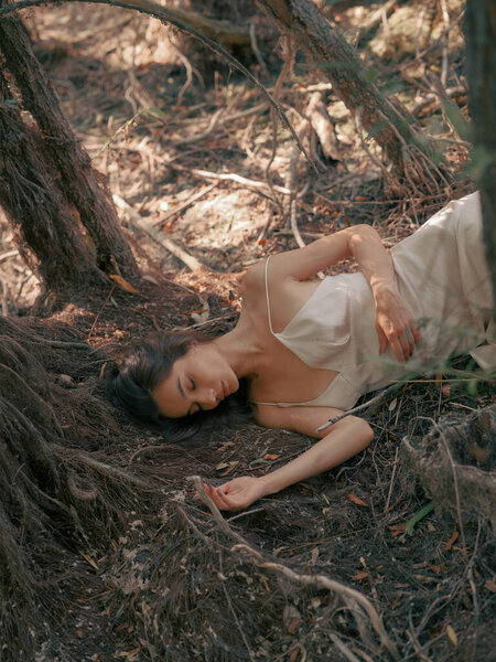 Young brunette woman in white dress lying down on ground and relaxing with closed eyes in forest, surrounded by old tree roots.Tranquil atmosphere, rest, relax and meditation in nature. Top view.