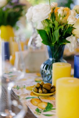 Salatalık turşusu, zeytin, mum ve zarif sofra takımlarıyla süslenmiş şık bir yemek masası. Çiçekli süs, kırsal ama modern tasarım. Yaz kutlaması ya da düğün ziyafeti. Yakın plan..