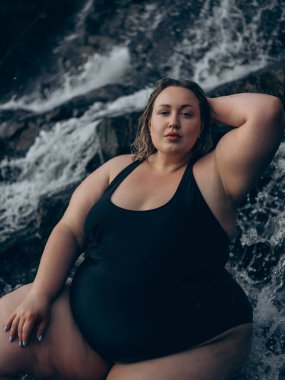 Siyah mayolu, kendinden emin, iri yarı bir kadın şelalenin yanında kayaların üzerinde dinleniyor yaz güneşinin tadını çıkarıyor, dışarıda vücut pozitifliğini ve doğal güzelliği kucaklıyor. Yakın plan. Portre.