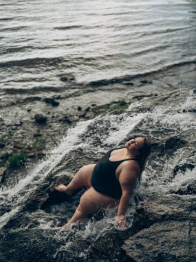 Siyah mayolu, kendinden emin, iri yarı bir kadın şelalenin yanında kayaların üzerinde dinleniyor yaz güneşinin tadını çıkarıyor, dışarıda vücut pozitifliğini ve doğal güzelliği kucaklıyor. Üst görünüm.