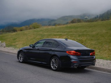 Parlak siyah sedan yola park edilmiş, modern tasarım sergileniyor, parlak dış görünüş, şık detaylar ve güçlü bir varlık. Lüks araç zarafeti, hızı ve karmaşıklığı simgeliyor..