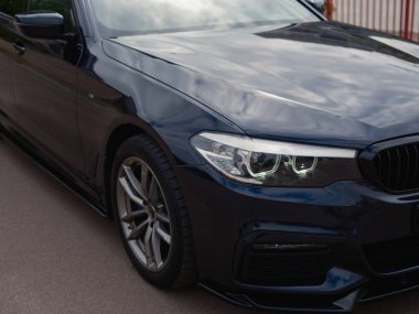 Modern koyu renkli sedanın ön tarafını gösteren yakın plan fotoğraf. Parlak far, şık alaşımlı tekerlek ve parlak boyaya odaklan. Otomotiv tasarımı ve teknoloji temaları için ideal.