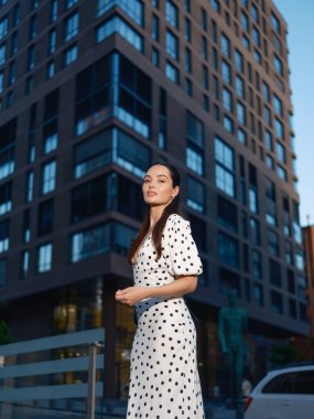 Puantiyeli genç, şık bir kadın akşam saatlerinde modern bir apartmanın önündeki bir şehir caddesinde kendinden emin bir şekilde duruyor..