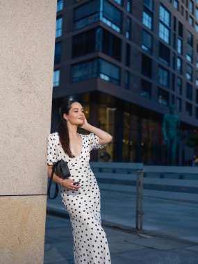 Puantiyeli elbisesiyle genç, şık bir kadın alacakaranlıkta taş bir sütuna yaslanıyor. Şık, modern şehir yaşam tarzı anı.