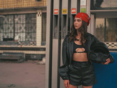 Deri ceketli ve bandanalı kendine güvenen genç motorcu kadın eski benzin pompalarının önünde motosiklet kaskı tutuyor. Şık, sert ve modern binici..