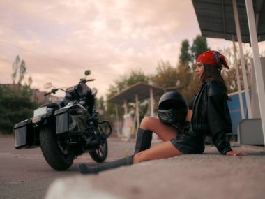 Deri ceketli genç, dalgın motorcu kadın ve kırmızı bandana eski benzin istasyonunun merdivenlerinde motosiklet kaskıyla oturuyor..