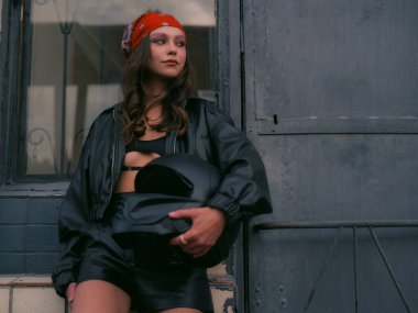 Kırmızı bandanalı, deri ceketli, kendine güvenen genç bir kadın şehir binasının yanında duran motosiklet kaskını tutuyor. Havalı, kendinden emin ve kentli motorcu tarzı..