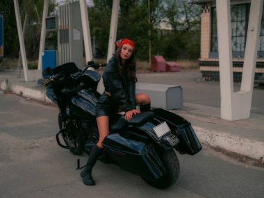 Siyah motosikletli güzel, kendine güvenen kadın motorcu, arkada oturuyor. Deri ceketli ve bandanalı şık genç bayan binici, şehir benzin istasyonu arkaplanı, özgürlük ve isyanı gösteriyor.