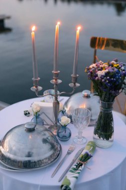 Mum ışığında romantik bir akşam yemeği ve gün batımında gölün arka planında bir buket çiçek. Özel etkinlik, yıldönümü, evlenme teklifi, kutlama, samimi romantik atmosfer, güzel yemekler..