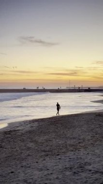 Deniz kıyısında yürüyen bir erkek sörfçünün silueti, gün batımı, dalgalar. Yüksek kalite 4k görüntü