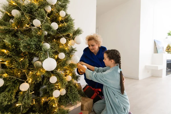 メリークリスマスとハッピーホリデー 陽気祖母と彼女のかわいい孫娘の女の子は贈り物を交換します おばあちゃんと小さな子供が屋内で木の近くで楽しんでいます プレゼント付きの家族を愛する — ストック写真
