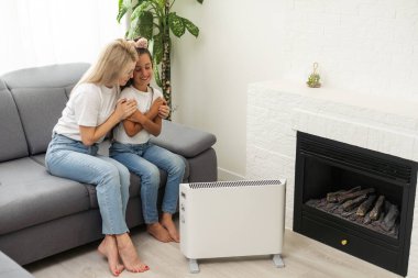 Anne ve çocuk evde elektrikli ısıtıcının yanında ellerini ısıtıyorlar..