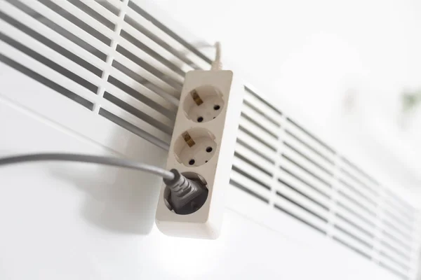 Electric heater plug inserted into extension cord. White radiator for home and office. House heating and comfort.