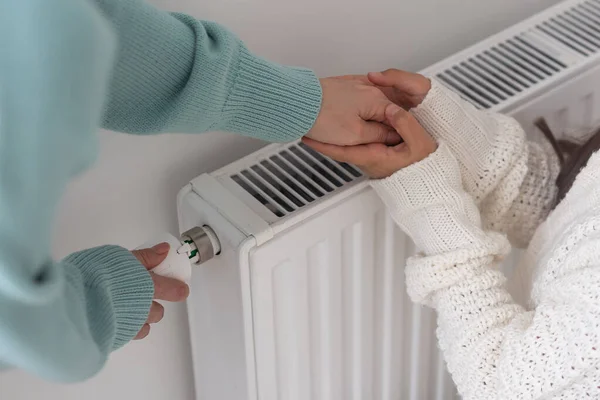 Child warming hands on heating radiator near white wall, closeup.