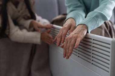 Elektrikli ısıtıcının başında, insanlar evlerinde sıcak bir battaniyeyle kendilerini ısıtıyorlar. Anne ve kızının elleri ısıtıcının yanında. Soğuk mevsim ve Avrupa 'da gaz krizi.