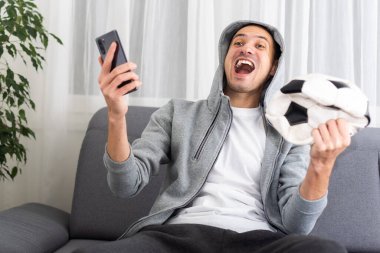 Şok olmuş futbol fanatiği mavi tişörtlü, akıllı telefon kullanan ve futbol topu tutan. Yüksek kalite fotoğraf