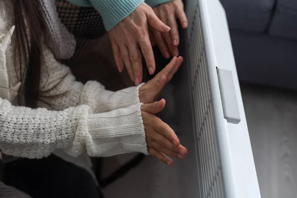 At the electric heater, people are warming themselves, covered with a warm blanket at home. hands of mother and daughter near the heater. Cold season and gas crisis in Europe.