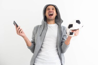 Şok olmuş futbol fanatiği mavi tişörtlü, akıllı telefon kullanan ve futbol topu tutan. Yüksek kalite fotoğraf