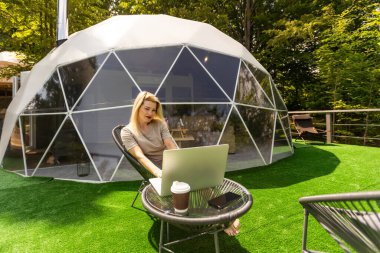 Dizüstü bilgisayar coğrafi çadırlarında çalışan bir kadın. Yeşil, mavi arka plan. Rahat, kamp, glamping, tatil, tatil yaşam tarzı konsepti. Açık hava kulübesi, manzaralı arka plan.