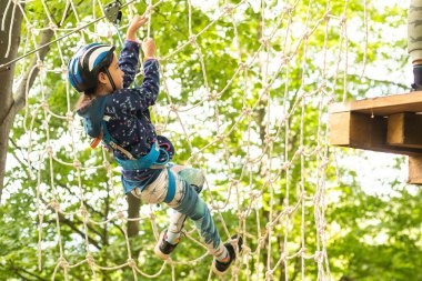 Little girl preschooler wearing full climbing harness having fun time in the rope park using carabiner and other safety equipment. Summer camp activity for kids. Adventure park in the forest.