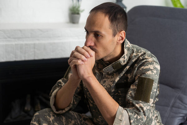 Thoughtful military man staring aside, holding palms by mouth, sitting on couch at home. Young soldier visiting psychologist, suffering from posttraumatic stress, closeup photo, copy space