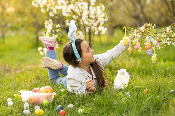 ウサギの耳の中で愛らしい女の子は 春の日に屋外で木の枝を咲かせます 庭でイースターエッグハントを楽しんでいる子供 — ストック写真