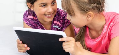 İki küçük tatlı kız dijital tablet kullanıyor ve çocuk odasında yatakta yatarken gülümsüyor..