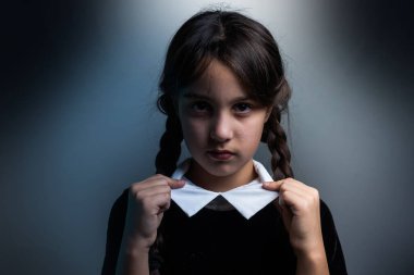 Cadılar Bayramı 'nda Çarşamba Addams kostümlü küçük bir kızın portresi. Ciddi ifade ve karanlık arkaplan ile karanlık atmosfer