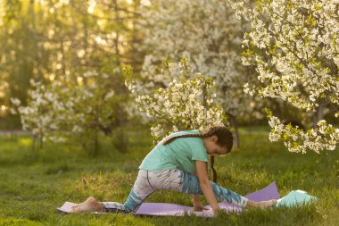 Küçük bir kız parkta bir spor minderinin üzerinde yoga yapıyor. Sağlıklı bir yaşam tarzı çocukça bir alışkanlıktır..