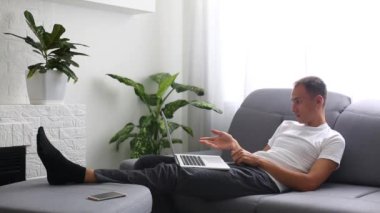 Evde kanepede çalışırken dizüstü bilgisayarının klavyesine bantlı kot gömlekli yakışıklı bir genç. Kapalı