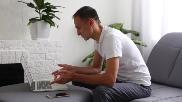 ソファの上で自宅で作業しながら ラップトップコンピュータのキーボードをタップジーンズシャツの若い良い外見の男 — ストック動画