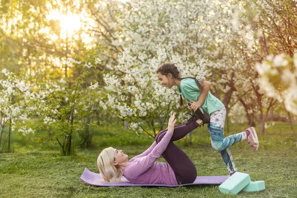Young sports mother doing physics exercise outdoors in garden near her daughter. Healthy lifestyle. Yoga. Fitness. Family having fun outside. Family spend time together in park