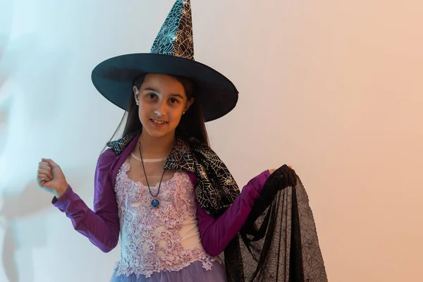 Happy scary Halloween concept: little witch girl playing paranormal Halloween role and doing scary look on a yellow background. Kid scaring and making Boo in carnival witch costume or wizard costume.