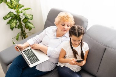 Yetişkin bir büyükanne, çocuğun ödevlerine evde yardım ediyor. Tatmin olmuş yaşlı büyükanne torununa oturma odasında ders çalışmasında yardım ediyor. Küçük kız yanında öğretmeniyle birlikte deftere yazı yazıyor.