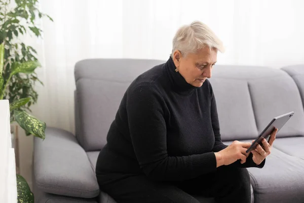 Smiling middle-aged Caucasian woman sit on couch in living room browsing wireless Internet on tablet, happy modern senior female relax on sofa at home using pad device, elderly technology concept.