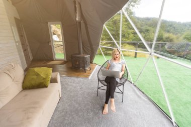 Kubbe kampında dizüstü bilgisayar kullanarak internette sohbet eden mutlu genç bir kadın. Glamping tatil konsepti.