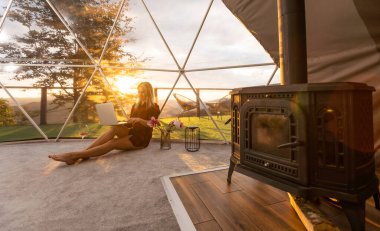 Kubbe kampında dizüstü bilgisayar kullanarak internette sohbet eden mutlu genç bir kadın. Glamping tatil konsepti.