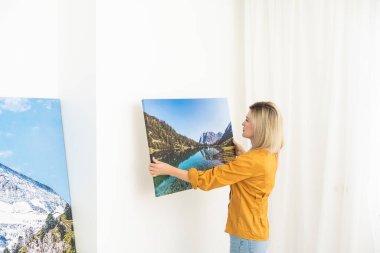 Canvas print with gallery wrap interior decor. Woman hangs landscape photography on white wall. Hands holding photo canvas print.