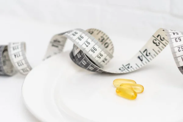 Dietary supplement for well-being. Fish oil or omega-3 capsules on plate near measuring tape on white background top view.