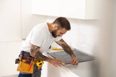 Adam mutfak yüzey dairesini elektrikle tamir ediyor. Tezgaha hob kuruluyor. Adam yeni mutfak mobilyaları ve aletler almış. Elektrikçi ev aletleri kurma işlemi