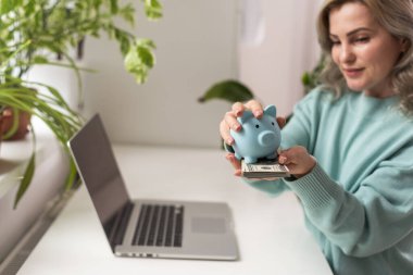 Para sevgisi. Gülümseyen kadın bir masada oturuyor ve birikimleriyle bir kumbarayı okşuyor. Başarılı ve kârlı online alışverişler yapan bir kadın gri arka planda bir masada oturuyor..