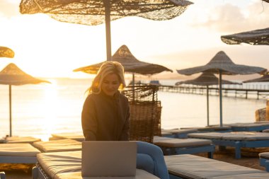 Kumsalda dizüstü bilgisayar kullanan genç bir kadın çalışıyor. Serbest çalışma, tatiller, mesafe çalışması, sosyal uzaklık, e-öğrenme, bağlantı, yaratıcı meslek, yeni iş, online konsept buluşması